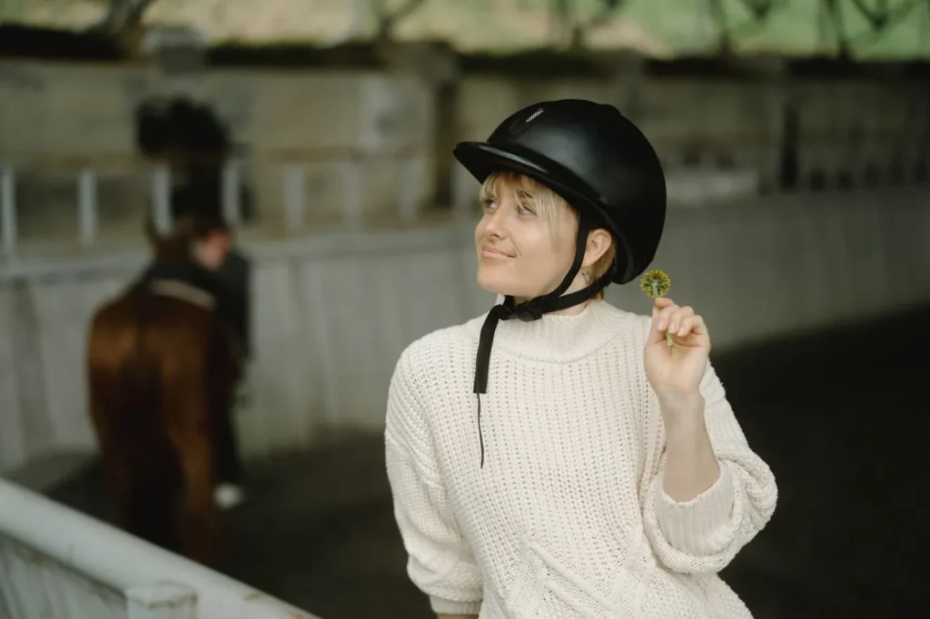 Rytter i tyk strikket sweater og sort ridehjelm med hagerem i en ridehal, med en hest i baggrunden